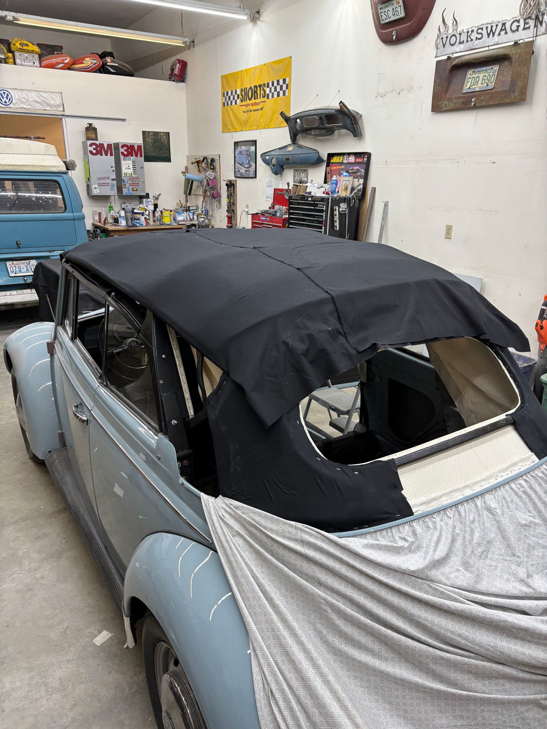Classic car in a garage with a black soft top and gray body panels, partially covered in protective fabric.