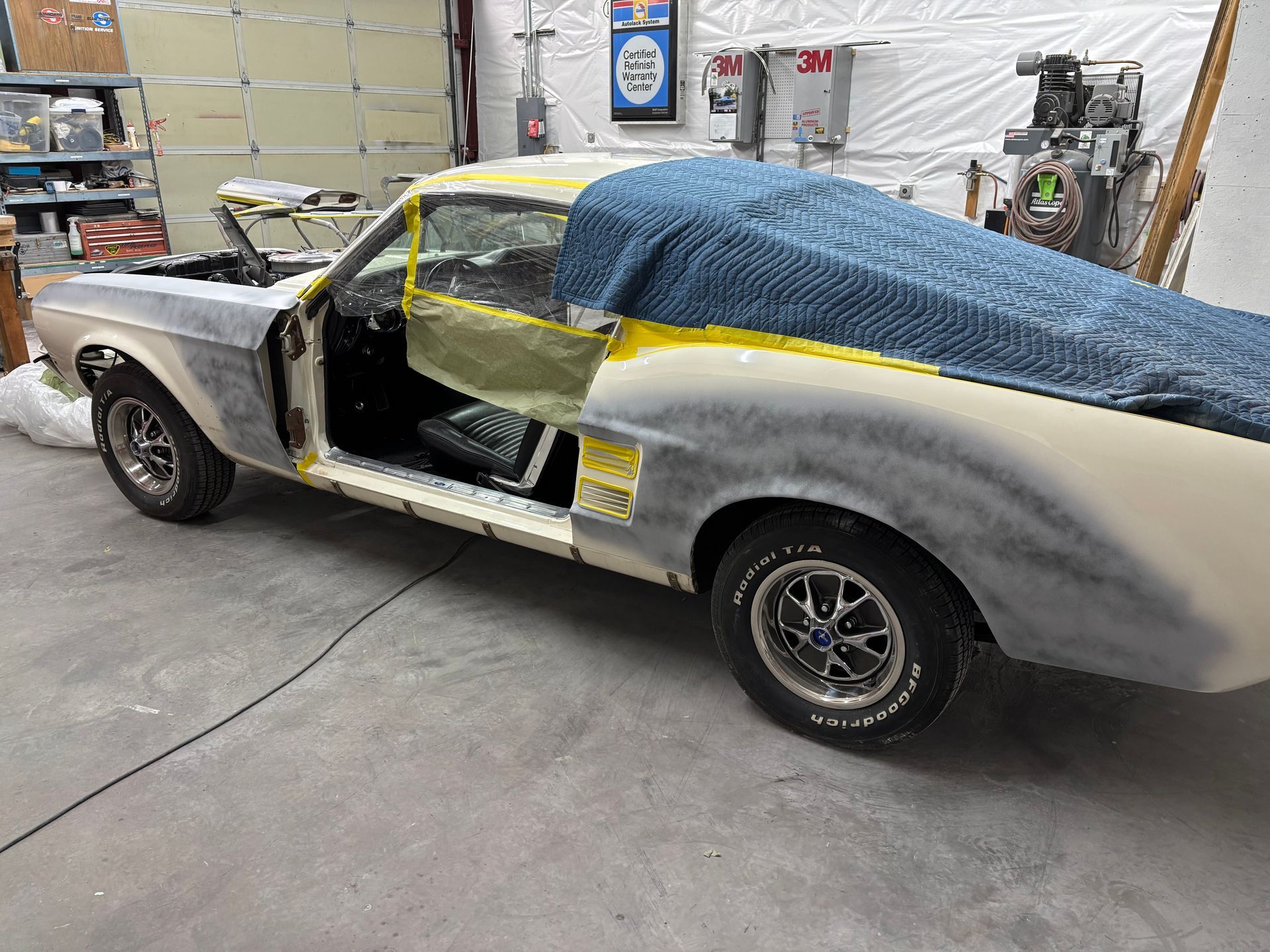 Classic car in a workshop, partially primed gray and masked with yellow tape and blue cover.