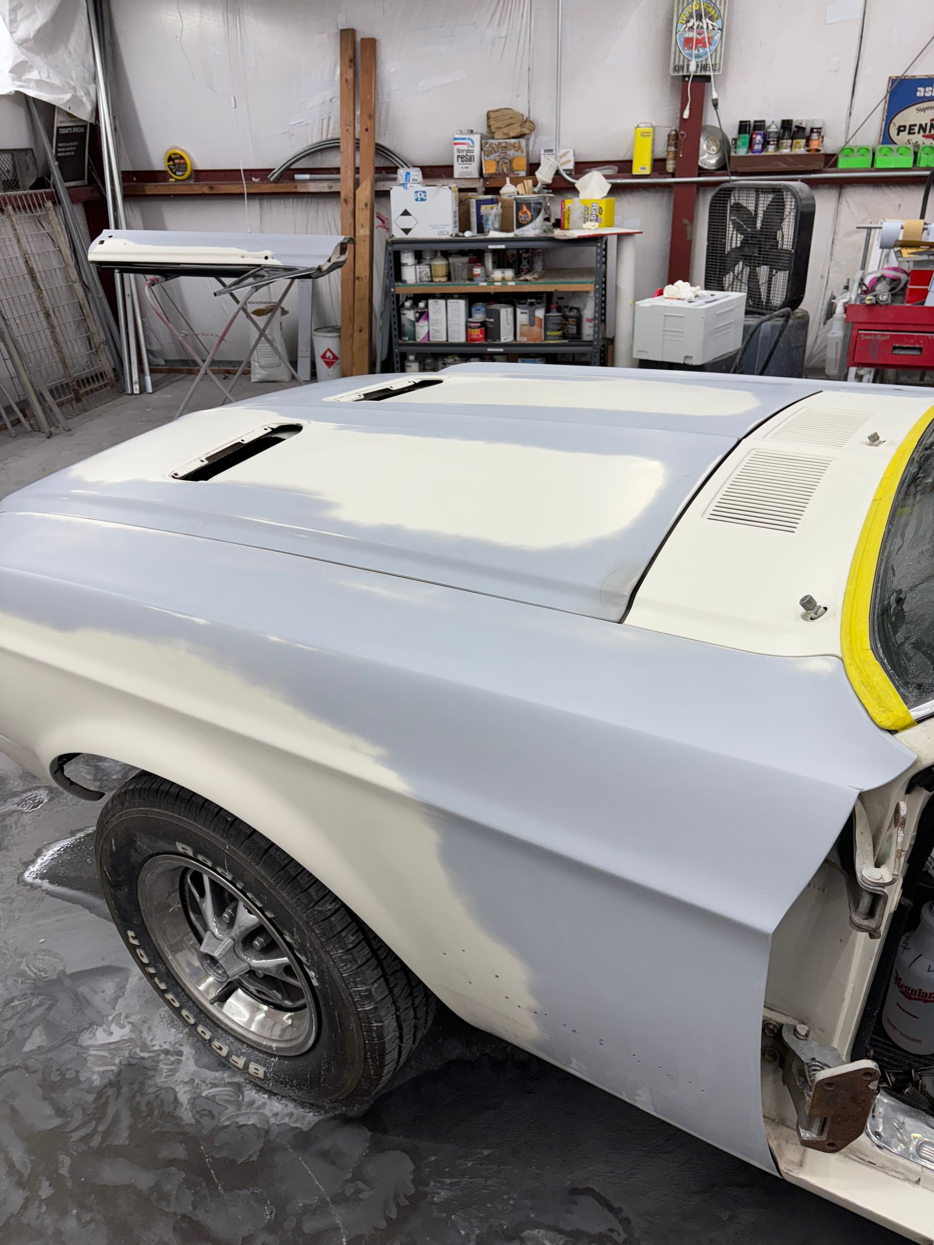 Primered classic car body in a garage, with a yellow-taped windshield area and rear wheel visible.
