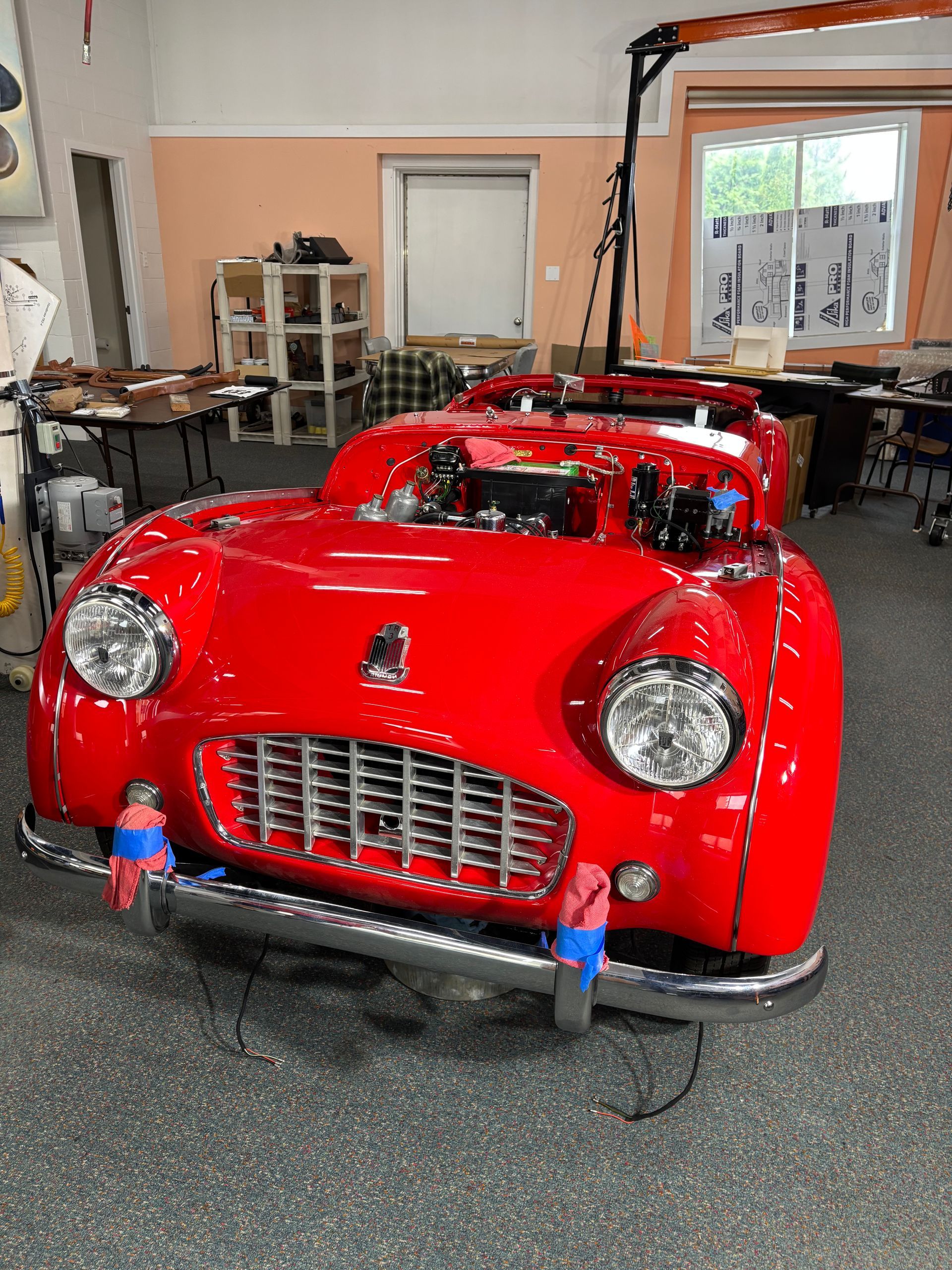 Red vintage convertible sports car indoors in a garage, front view with chrome grille and headlights.