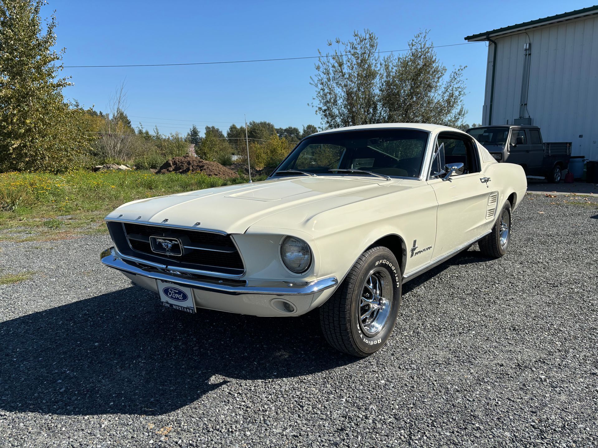 Cream-colored classic Ford Mustang parked on gravel near a building under a clear blue sky