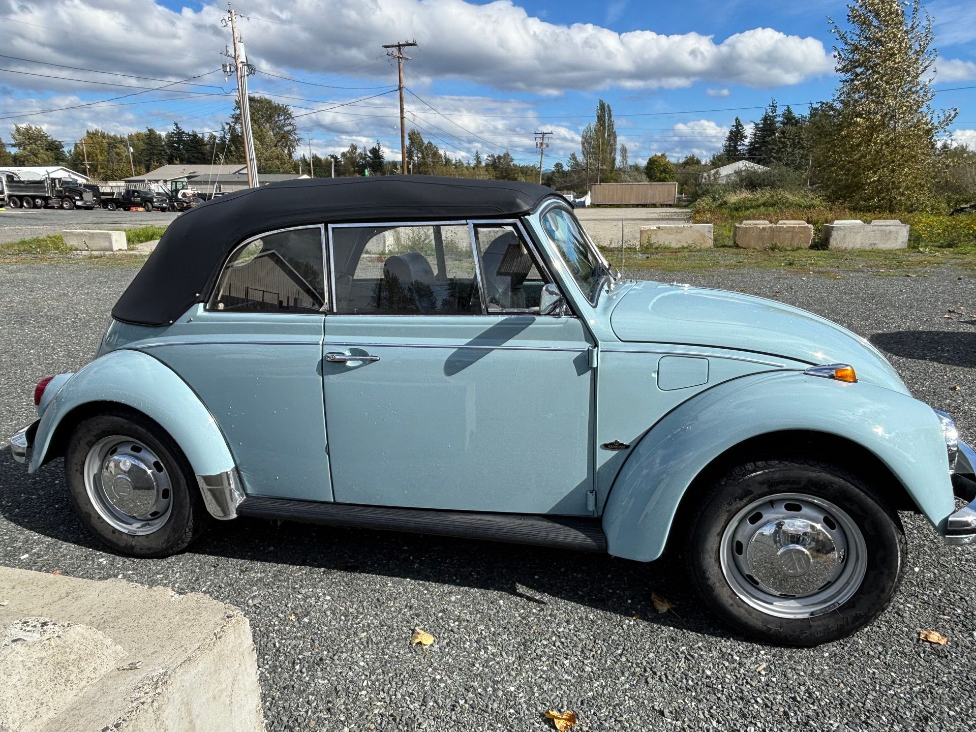 Light blue vintage Volkswagen Beetle with black convertible top parked on gravel under a cloudy sky