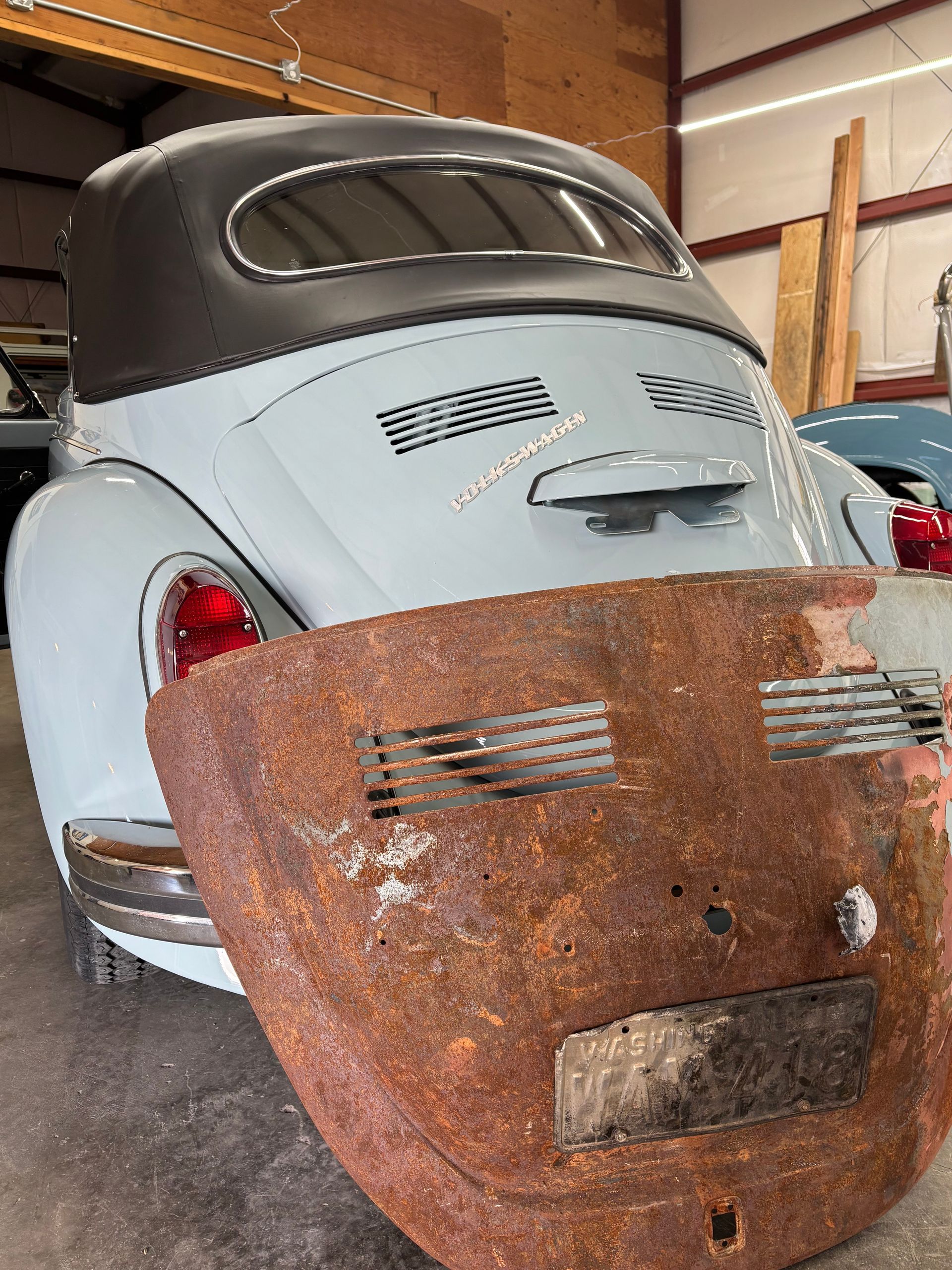 Rusty car body panels leaning in a garage, with a light blue vintage Volkswagen Beetle behind them.
