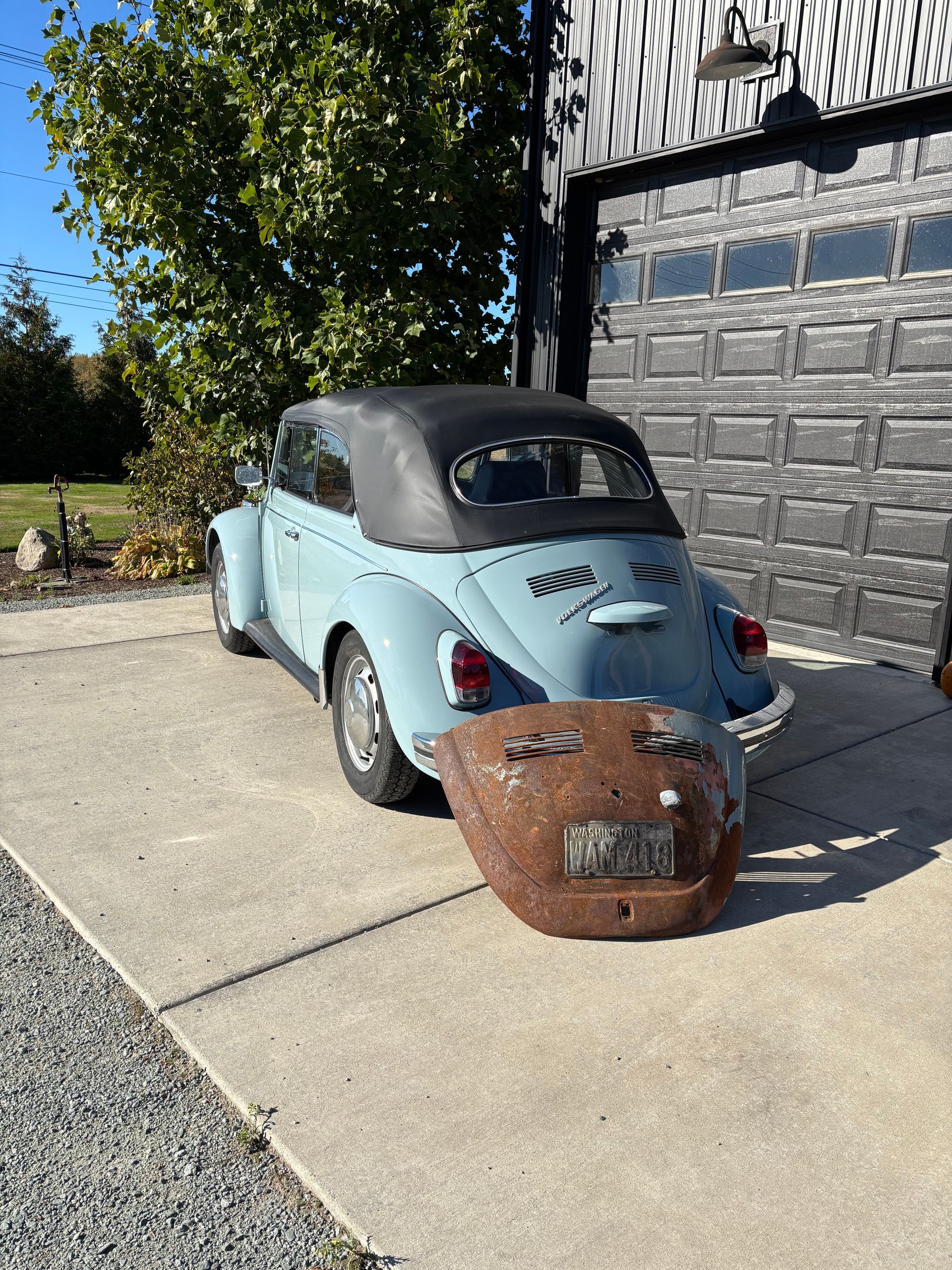 Vintage light blue convertible with a black top parked in a driveway beside a garage, with a rusty metal bin in front.