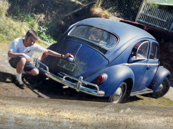 Man washing a blue Volkswagen Beetle in a driveway with a hose