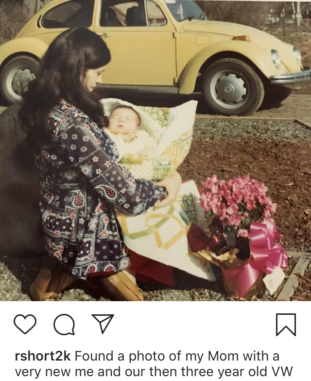 Woman in patterned dress holds a wrapped baby near pink flowers, with a yellow Volkswagen Beetle behind them.