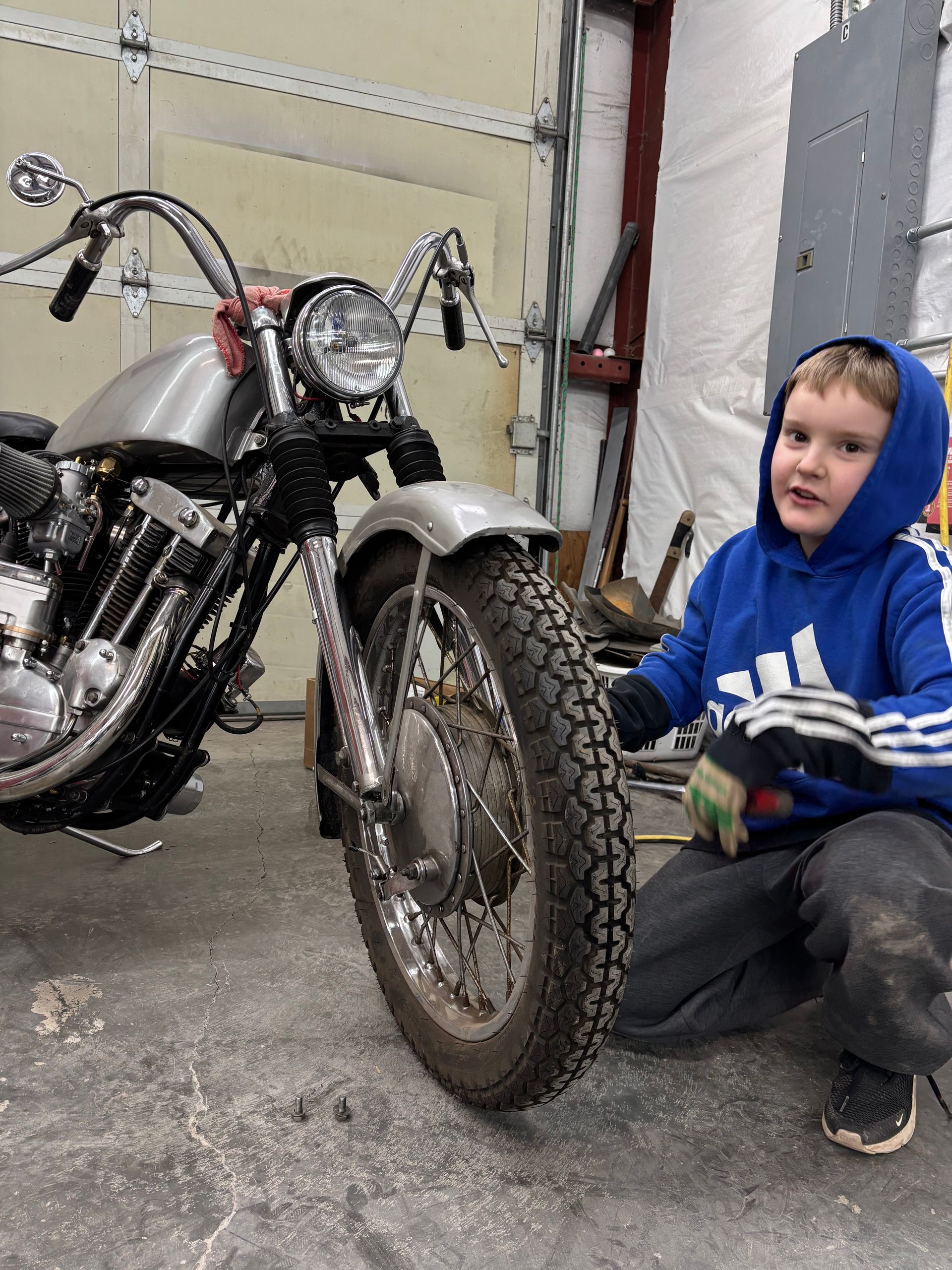 Child in blue hoodie crouches beside a motorcycle in a garage.