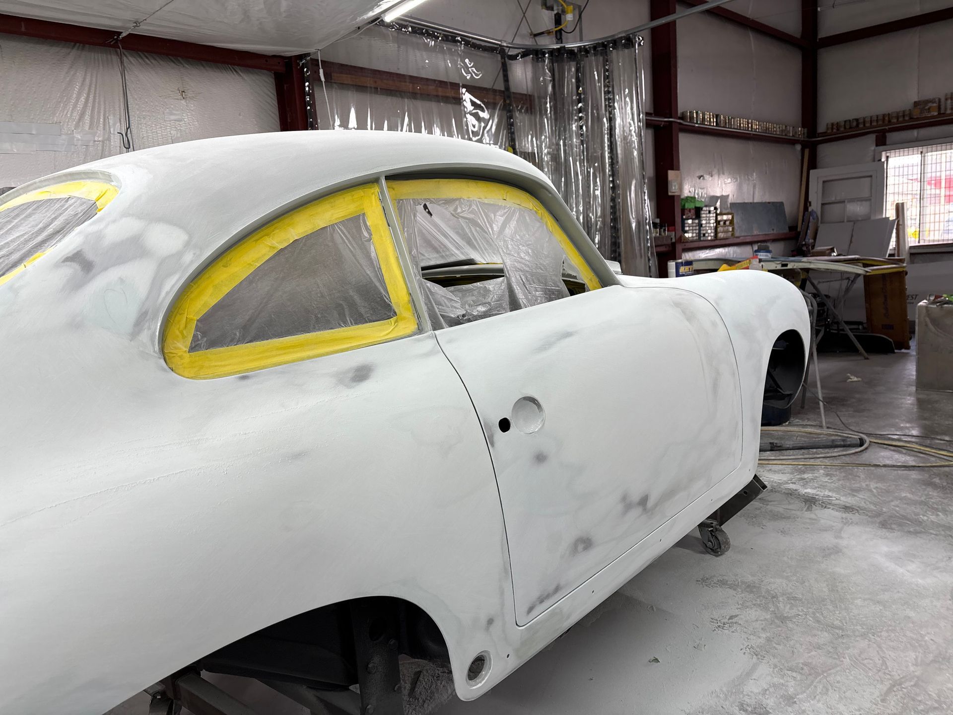Classic car body primed white in a workshop, with yellow-taped windows and a bare rear wheel opening
