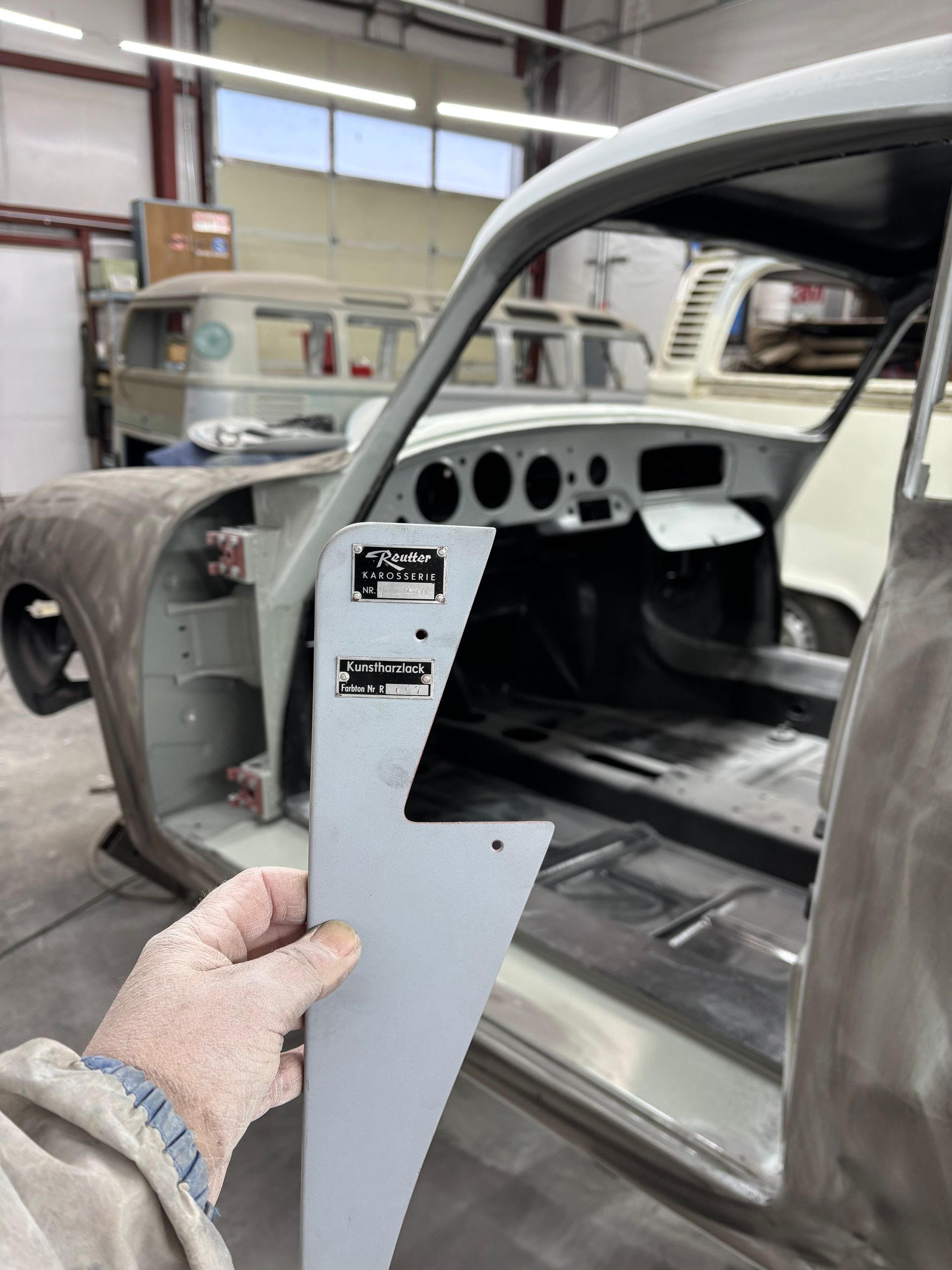 Hand holding a gray metal panel beside a partially stripped car body in a workshop