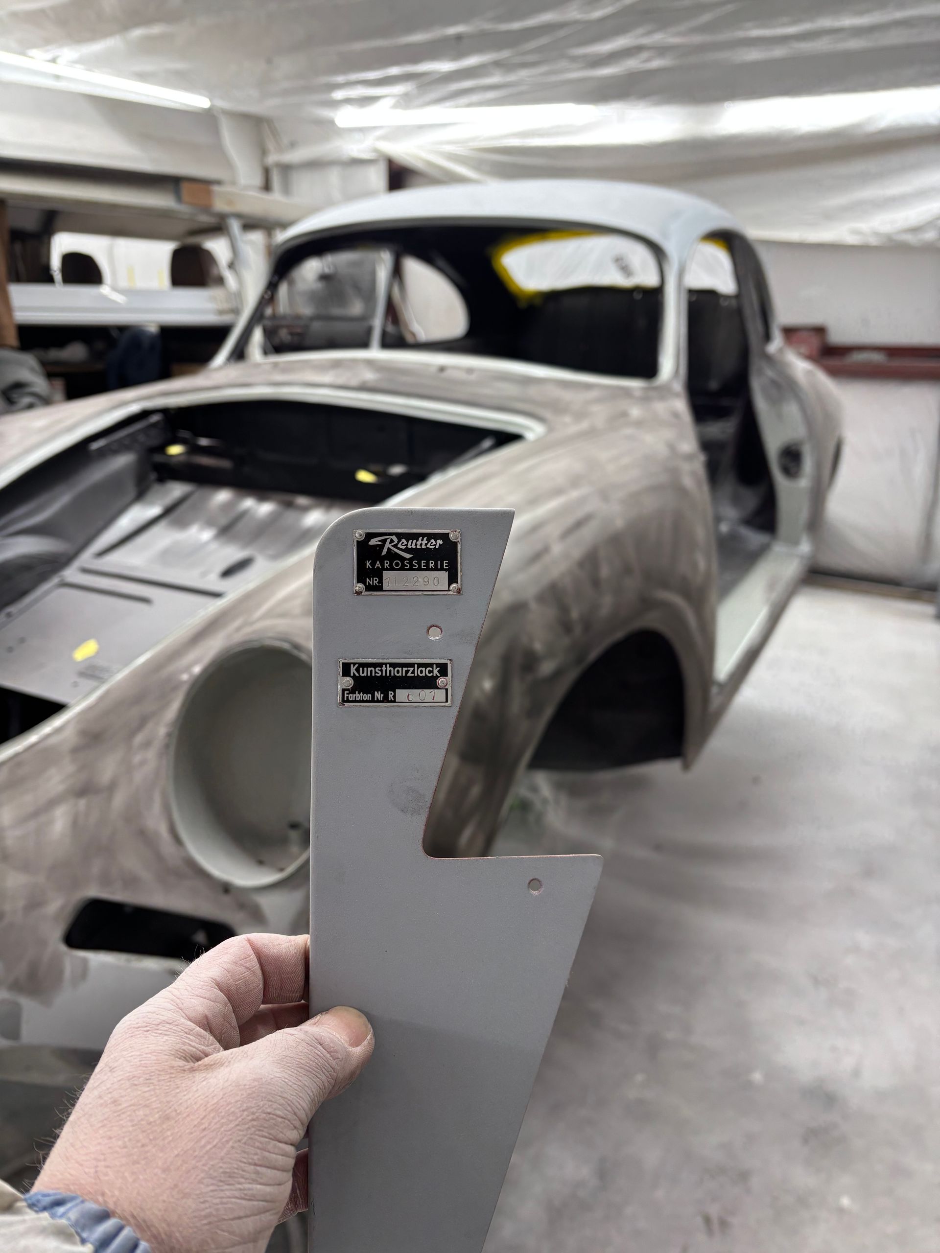 Hand holding a gray car body panel in front of an unfinished classic car in a workshop