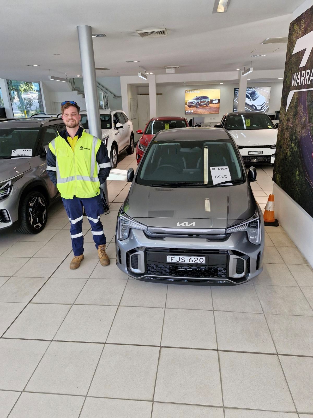 A man in a yellow vest is standing in front of a car in a showroom.