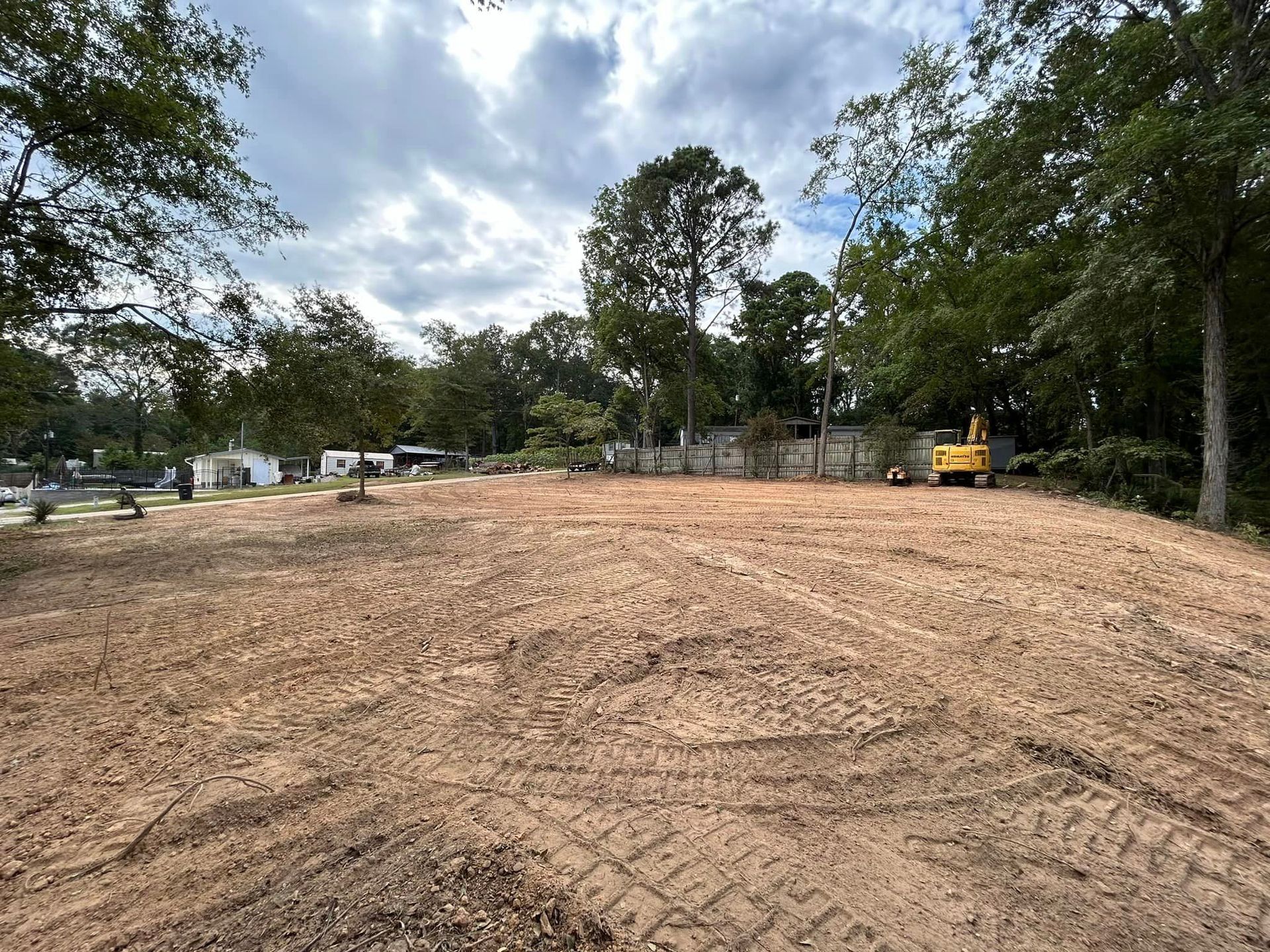 A large dirt field with trees in the background and a yellow excavator in the middle.