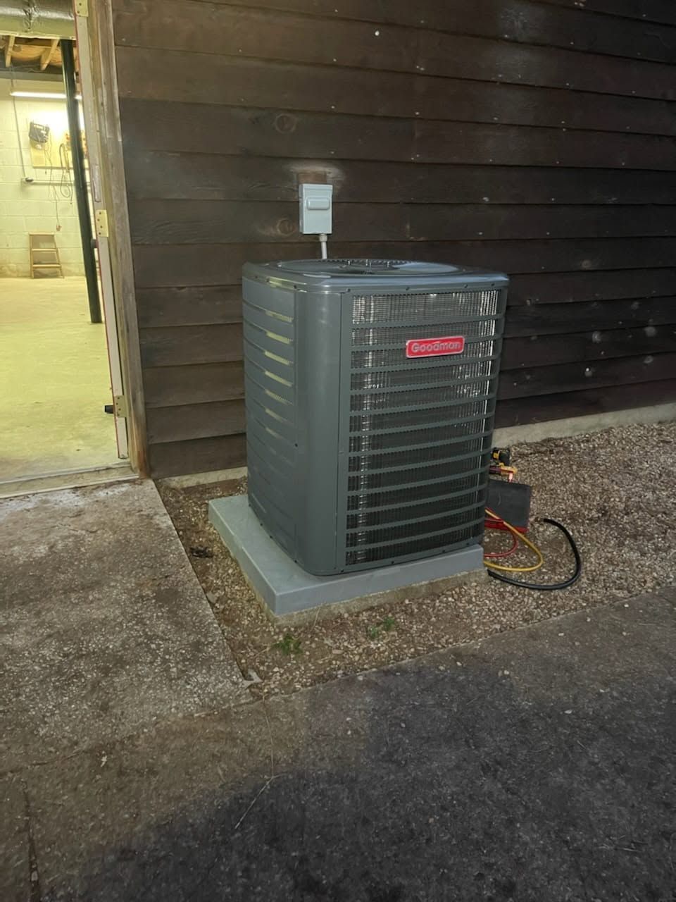 Outdoor air conditioner unit beside a brown-sided building, with a hose and concrete pad at dusk