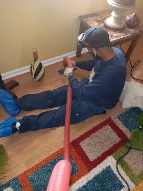 Person sitting on a hardwood floor holding a red vacuum hose in a living room.