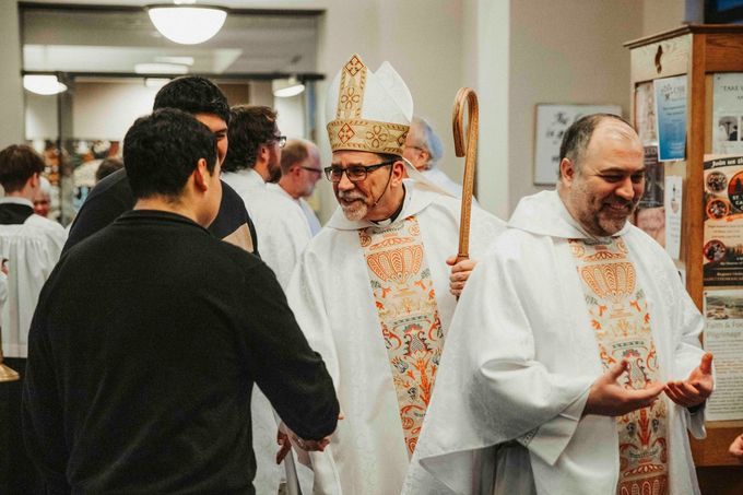 Clergy in white vestments greet each other in a hallway, one wearing a bishop’s mitre.