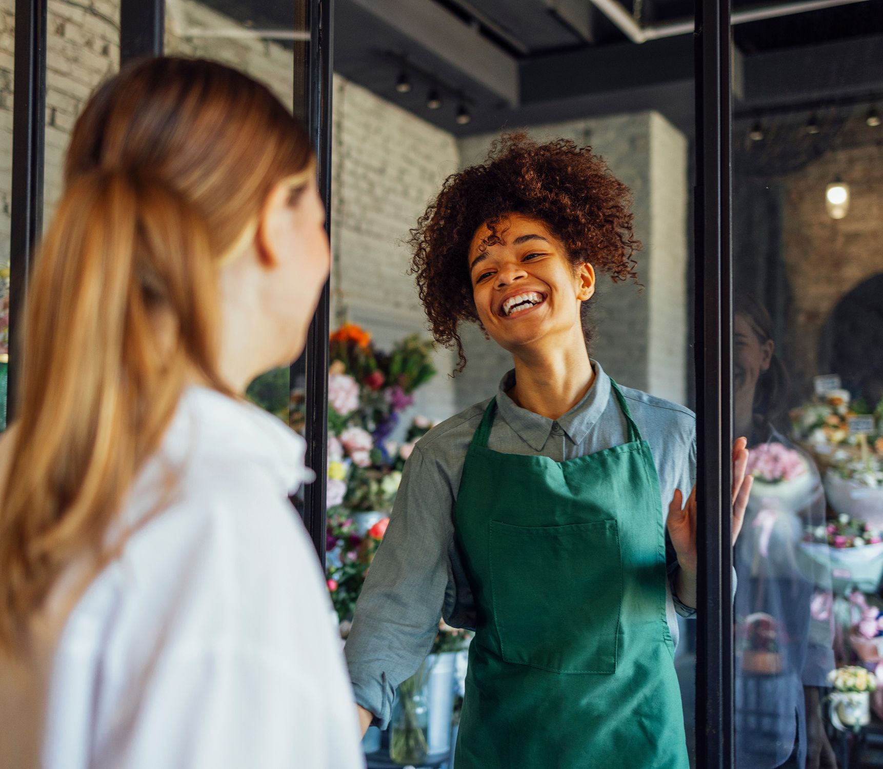 A woman in a green apron is talking to another woman in a flower shop.