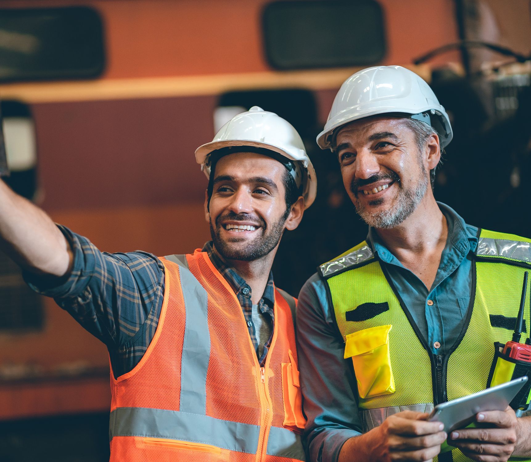 Two construction workers are standing next to each other and smiling while looking at a tablet.