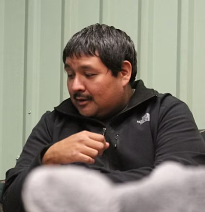 Man with dark hair in a black jacket, looking down, possibly indoors.