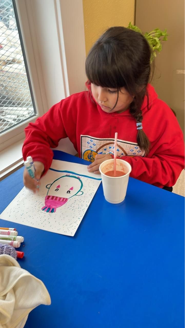 A young girl with a braid, drawing with markers at a blue table; a cup of red liquid sits beside her.