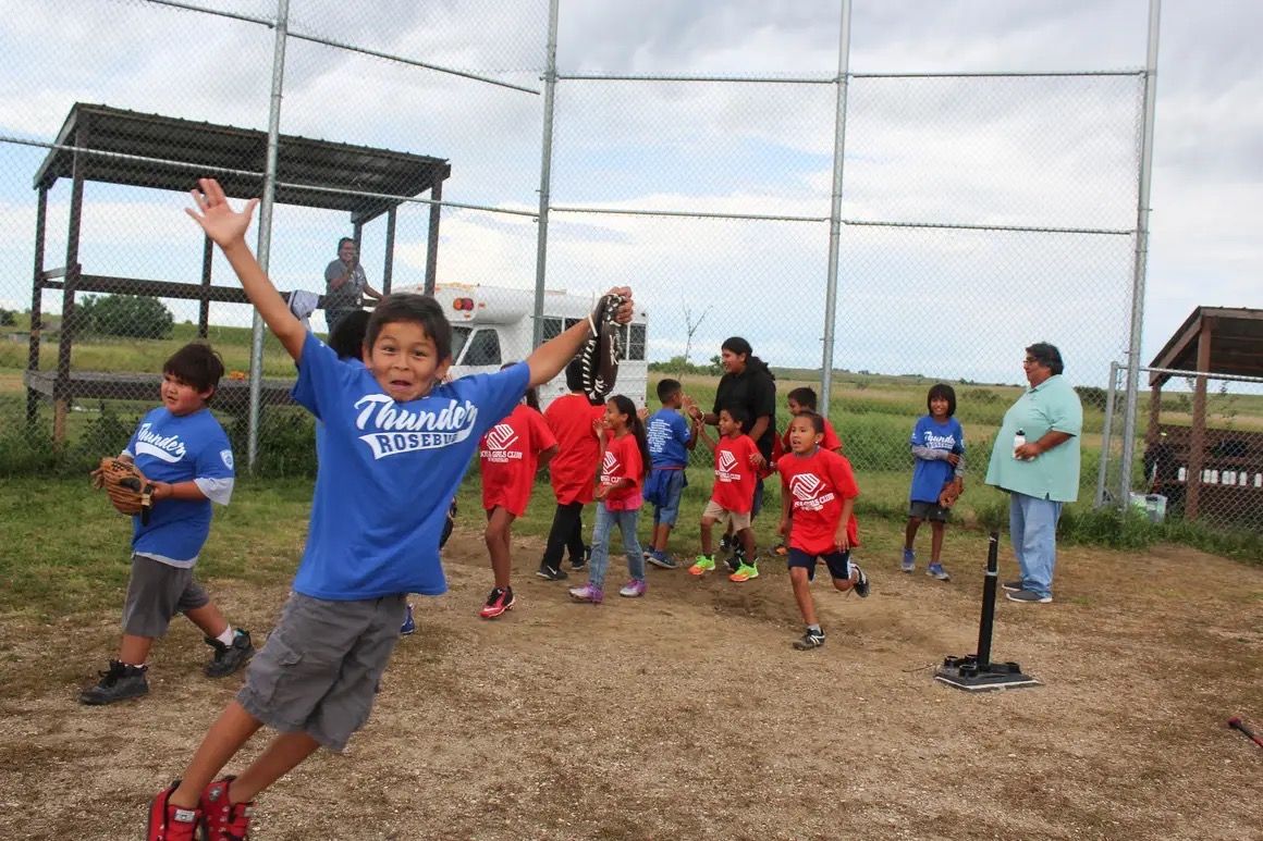 Children playing baseball on a dirt field; one boy jumps with arms raised, others run and stand.