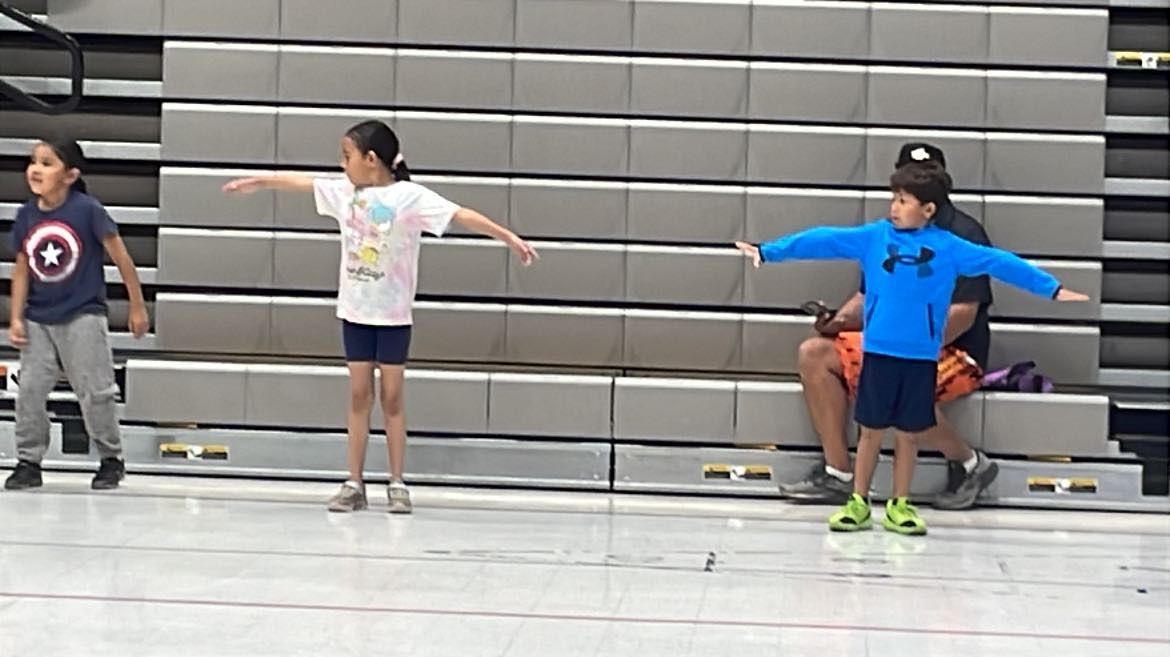 Three children with arms outstretched in a gym. A man sits behind them.