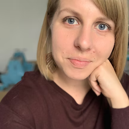 Woman with blonde hair, blue eyes, resting chin on hand, wearing a maroon top.