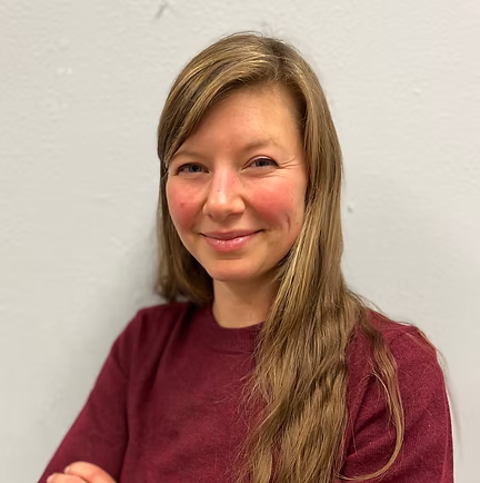 Woman with long brown hair and red sweater smiling, arms crossed, against a gray wall.