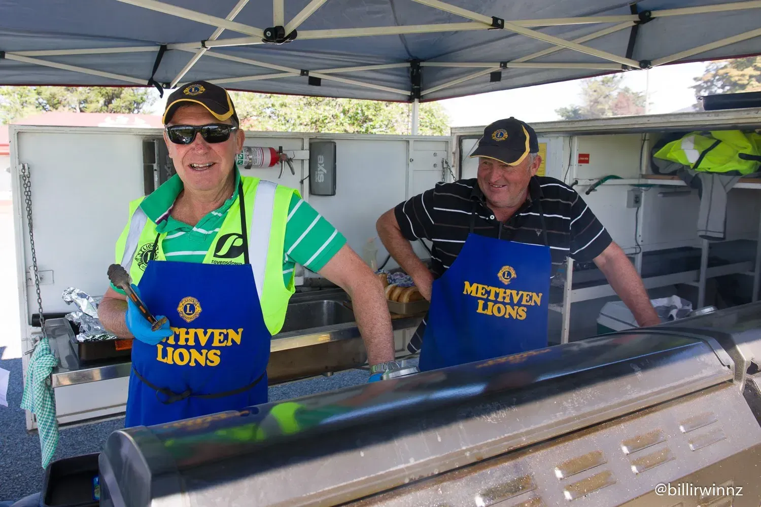Two men wearing blue aprons are standing in a kitchen.