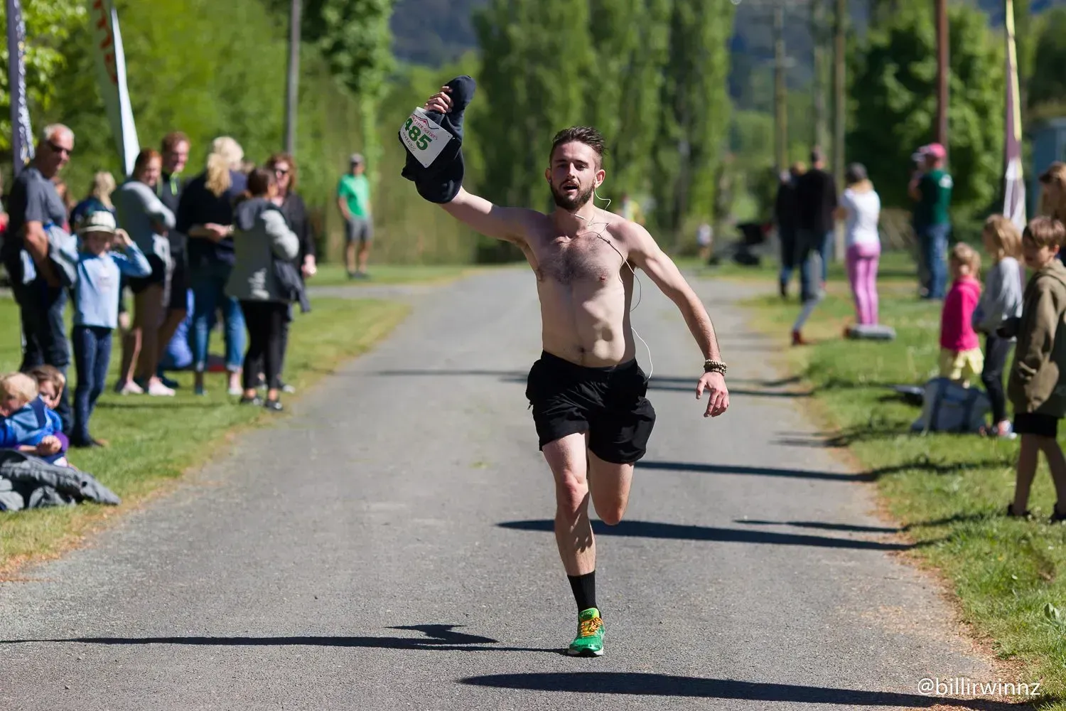 A shirtless man is running down a road while holding a bottle of wine in his hand.