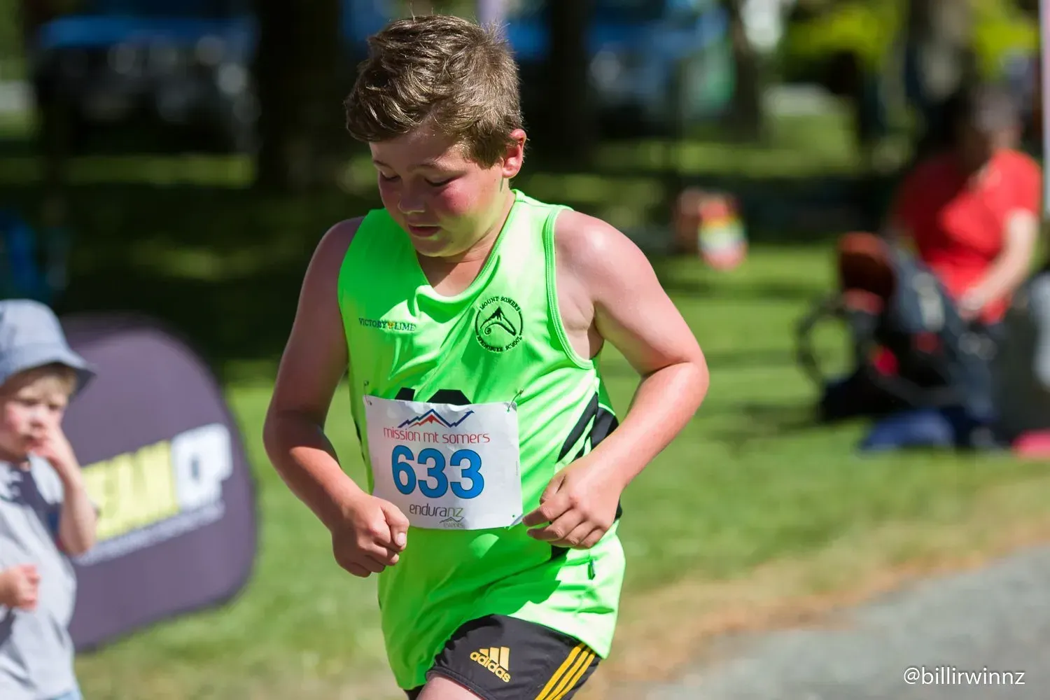 A young boy in a green tank top and shorts is running in a race.