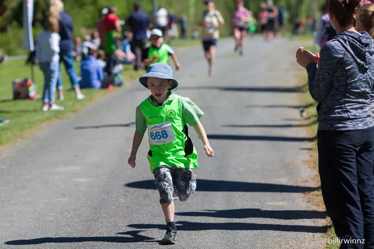 A young boy in a green shirt and hat is running down a road.