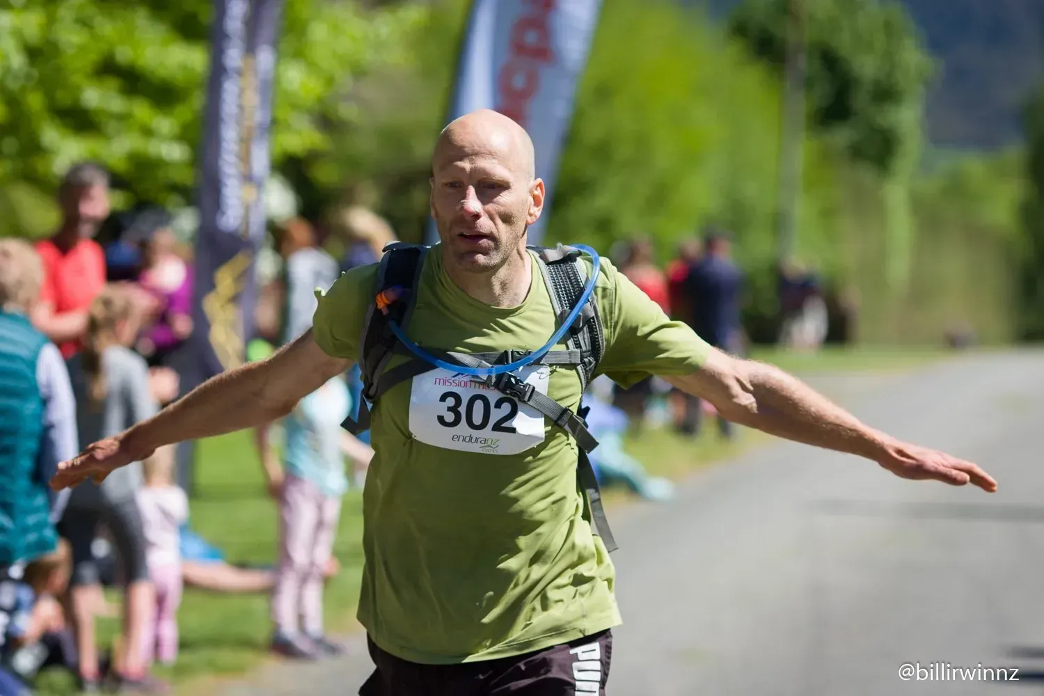 A man wearing a green shirt with the number 302 on it is standing on a road with his arms outstretched.