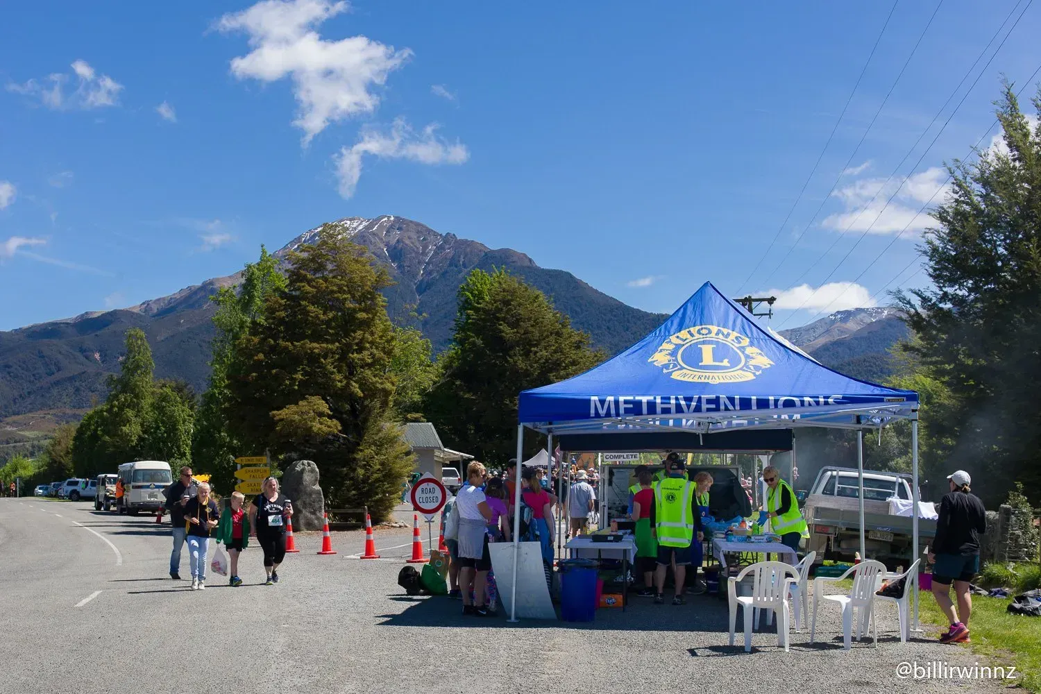 A group of people are standing under a blue tent with a mountain in the background.