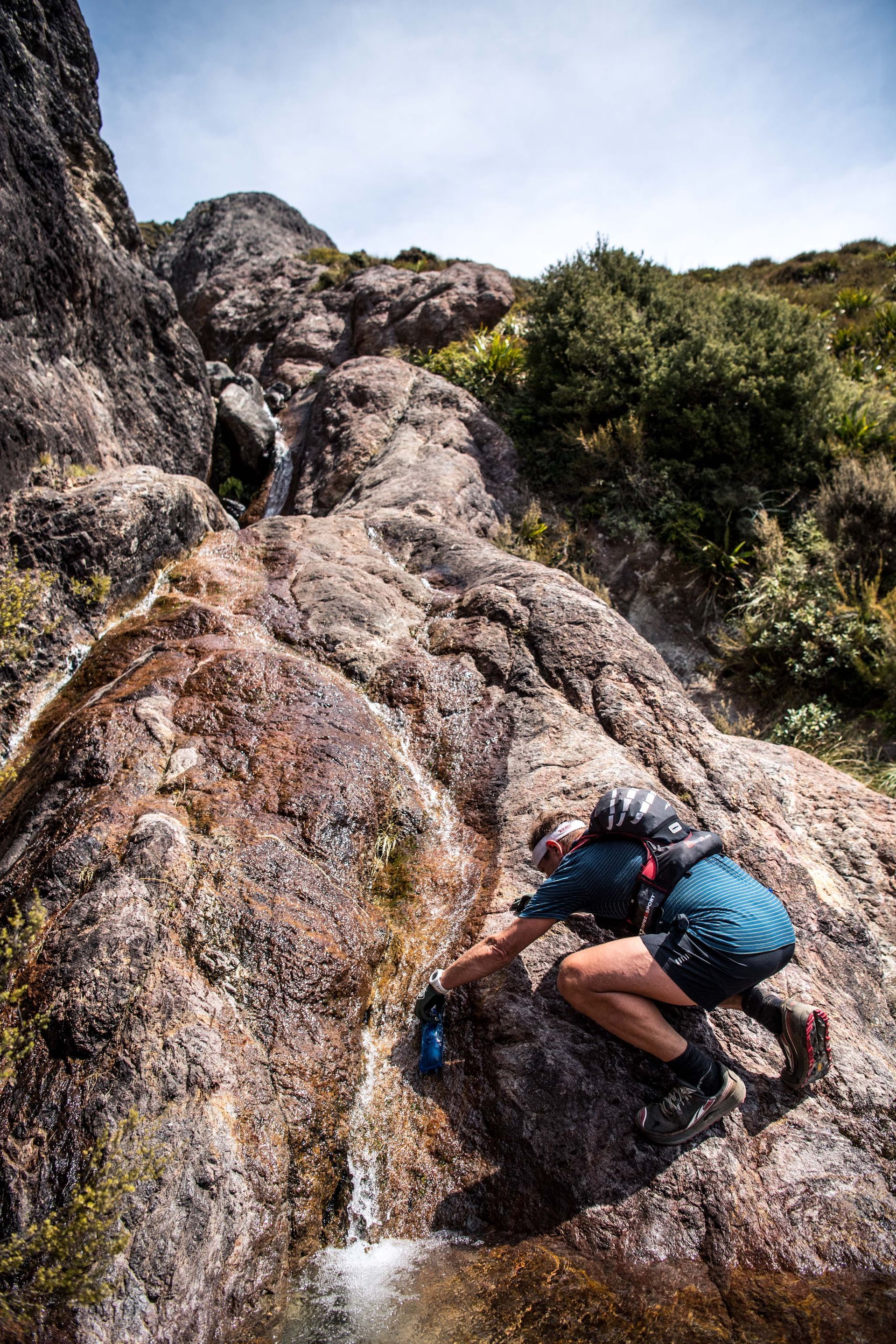 A man is drinking water from a small waterfall on a rock.