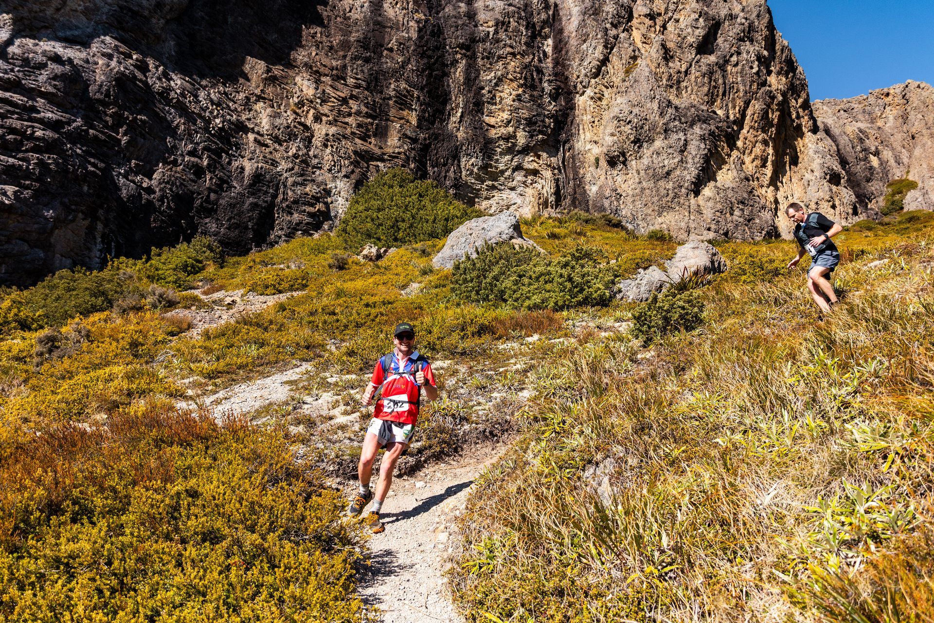 Two people are running down a trail in the mountains.