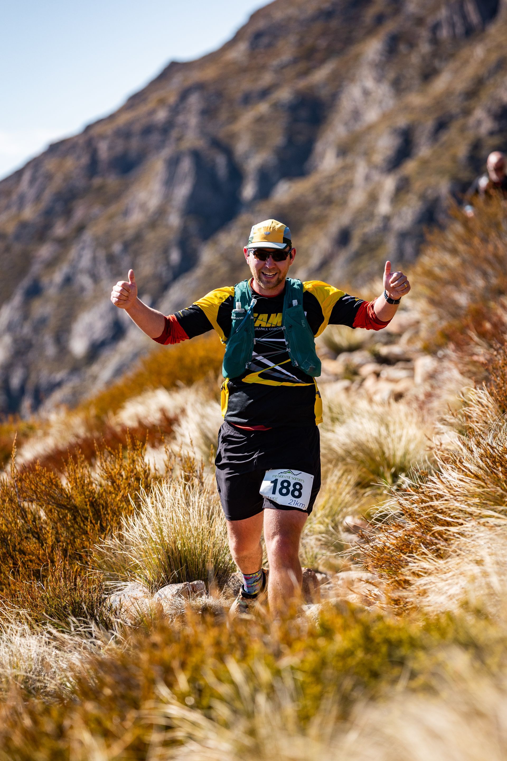A man is running on a trail in the mountains and giving a thumbs up.