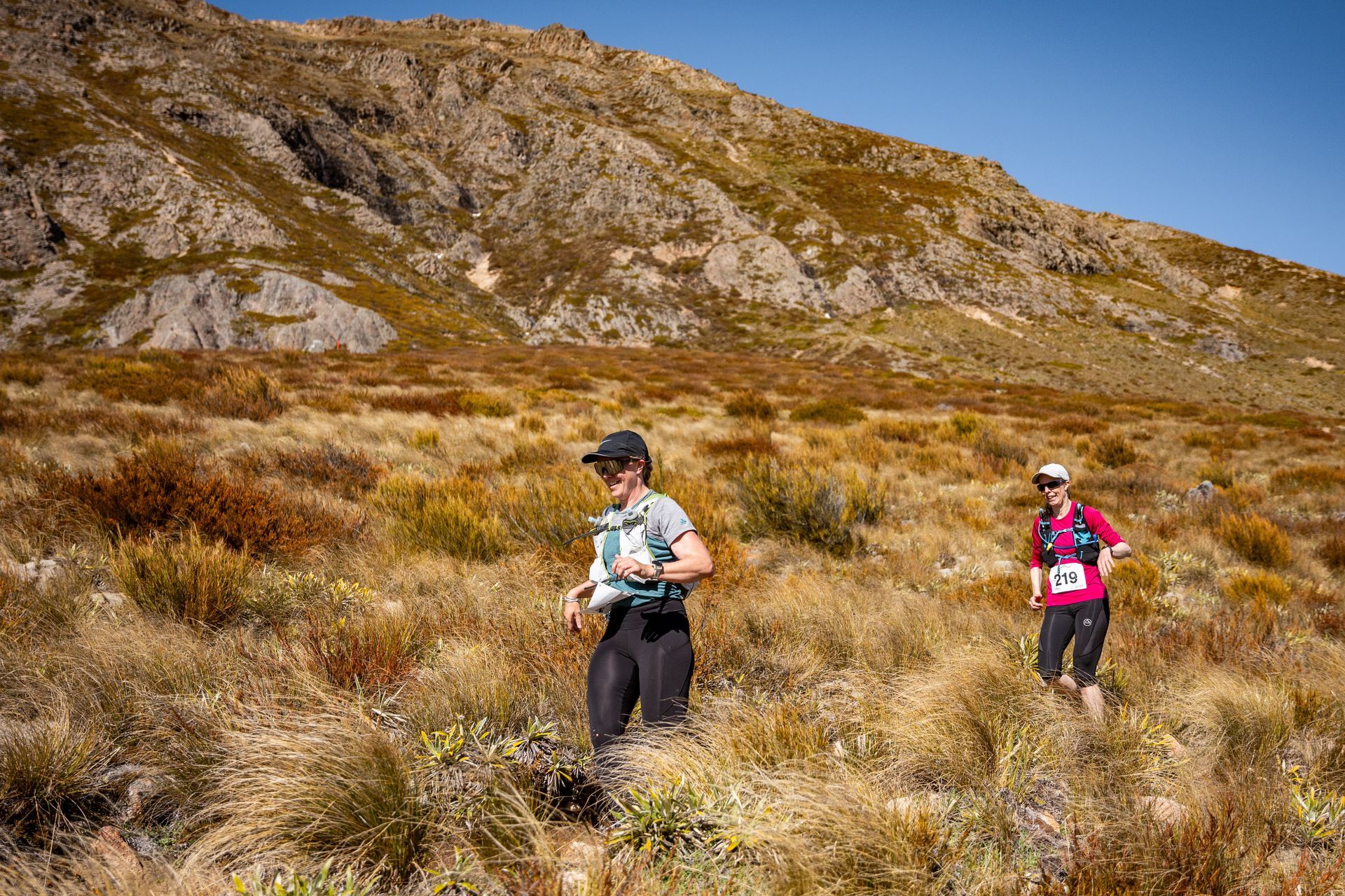 Two people are walking through a field with a mountain in the background.