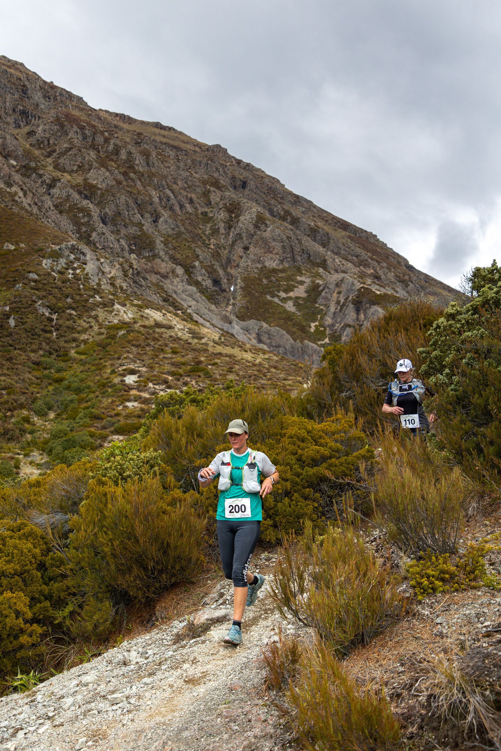 Two people are running down a trail in the mountains.