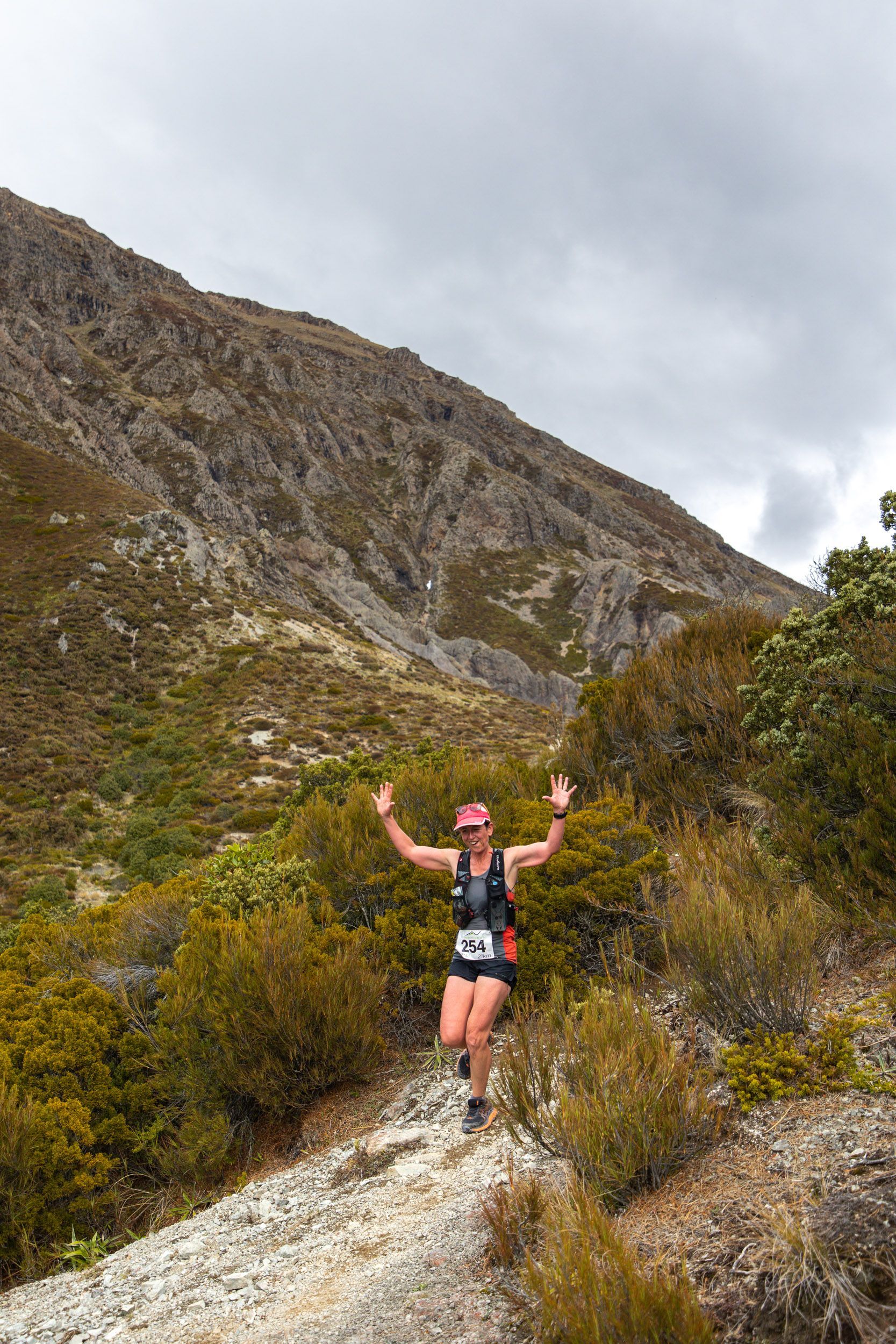 A woman is running on a trail in the mountains with her arms in the air.
