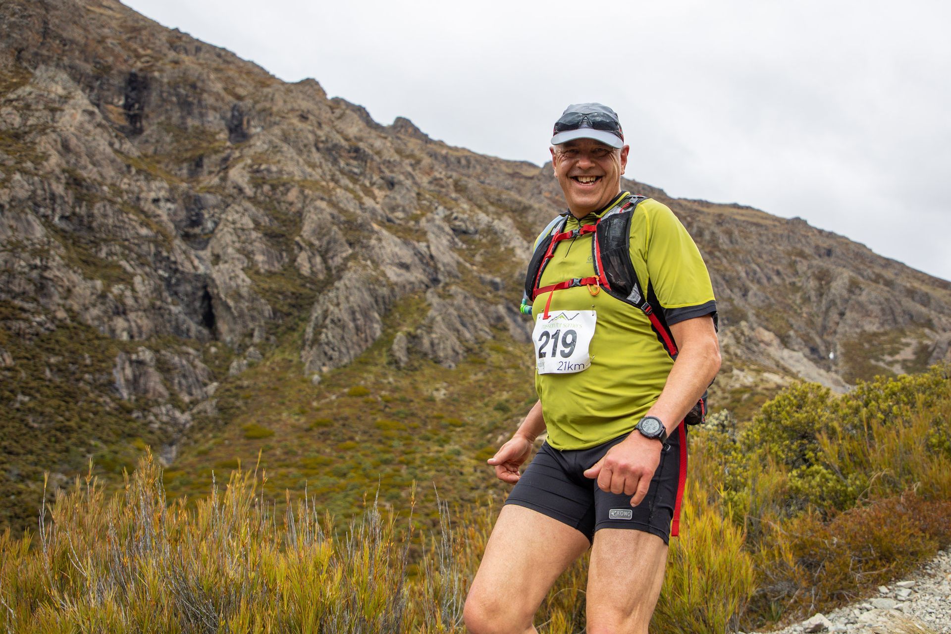 A man is standing in front of a mountain wearing shorts and a green shirt.
