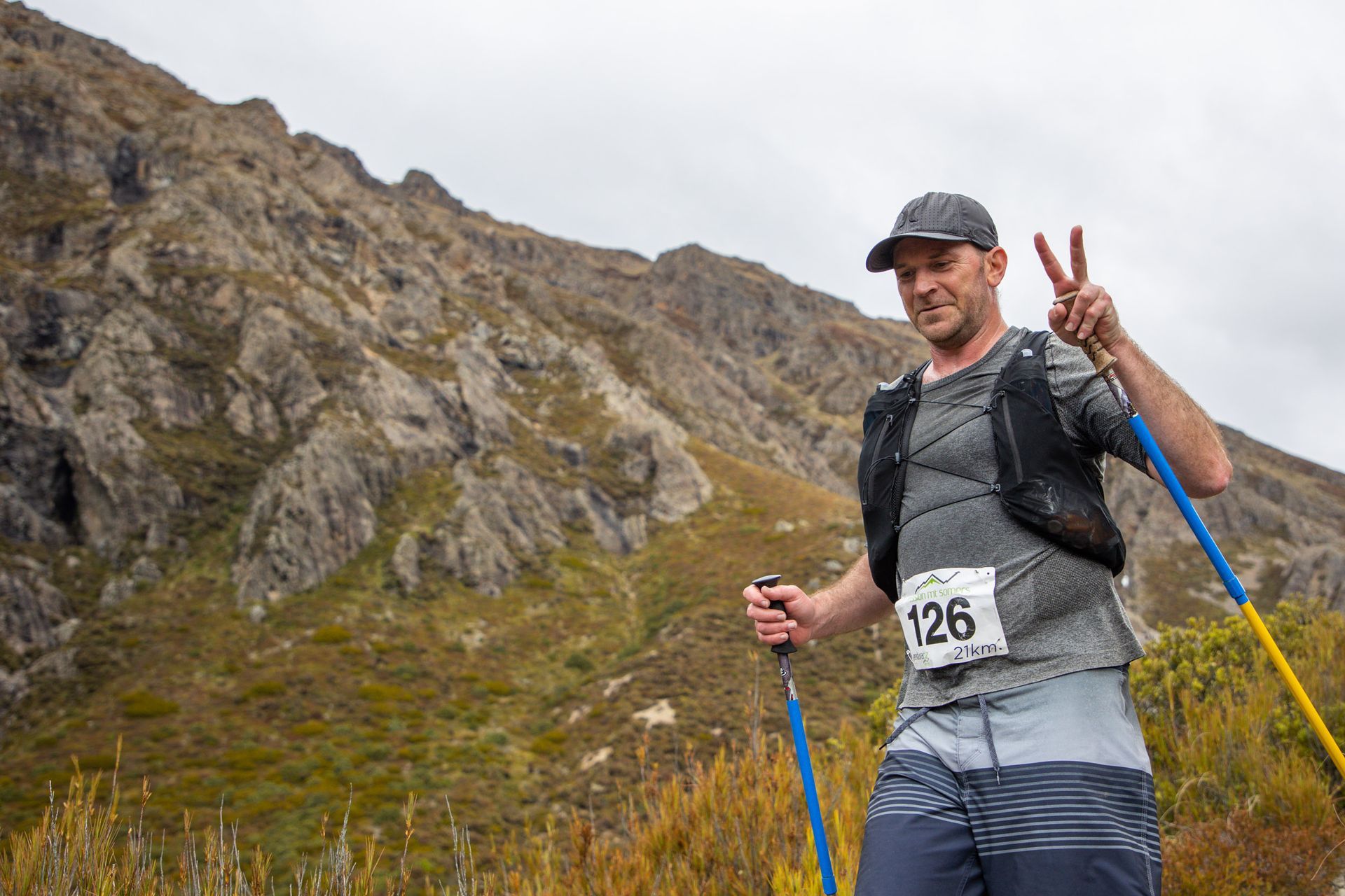A man is standing in front of a mountain holding hiking poles and giving a peace sign.