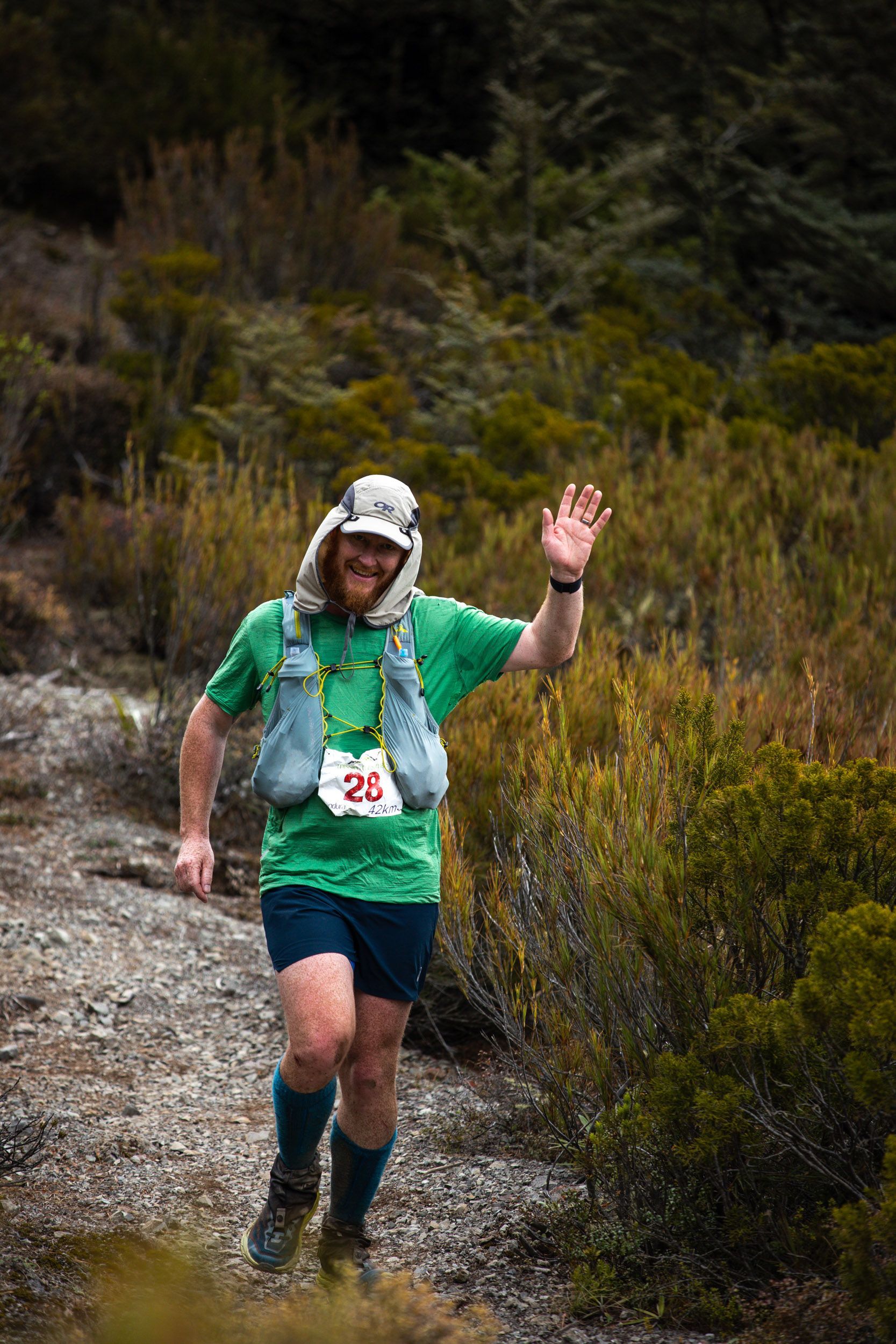 A man is running on a trail in the woods and waving at the camera.