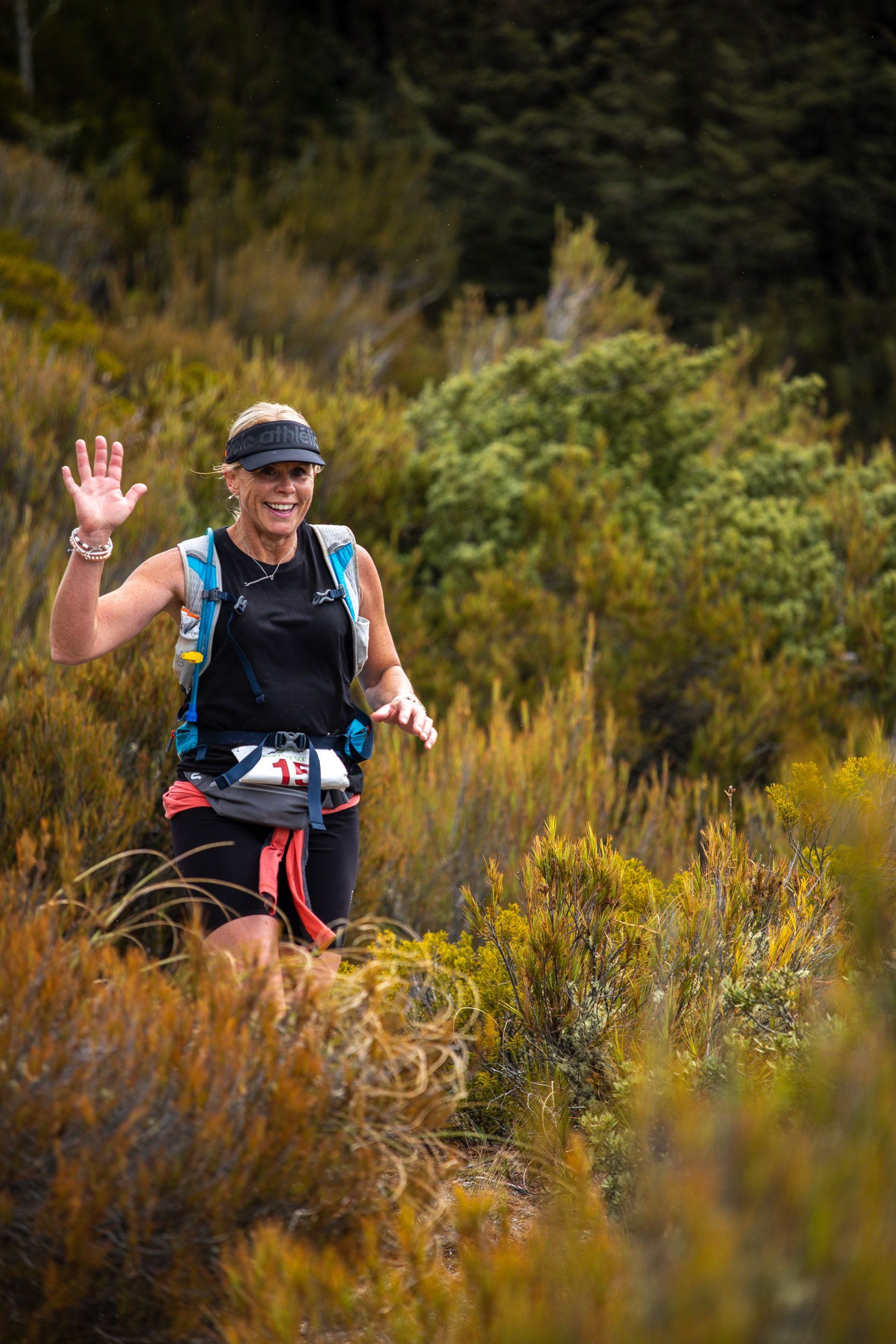 A woman is running on a trail in the woods and waving at the camera.