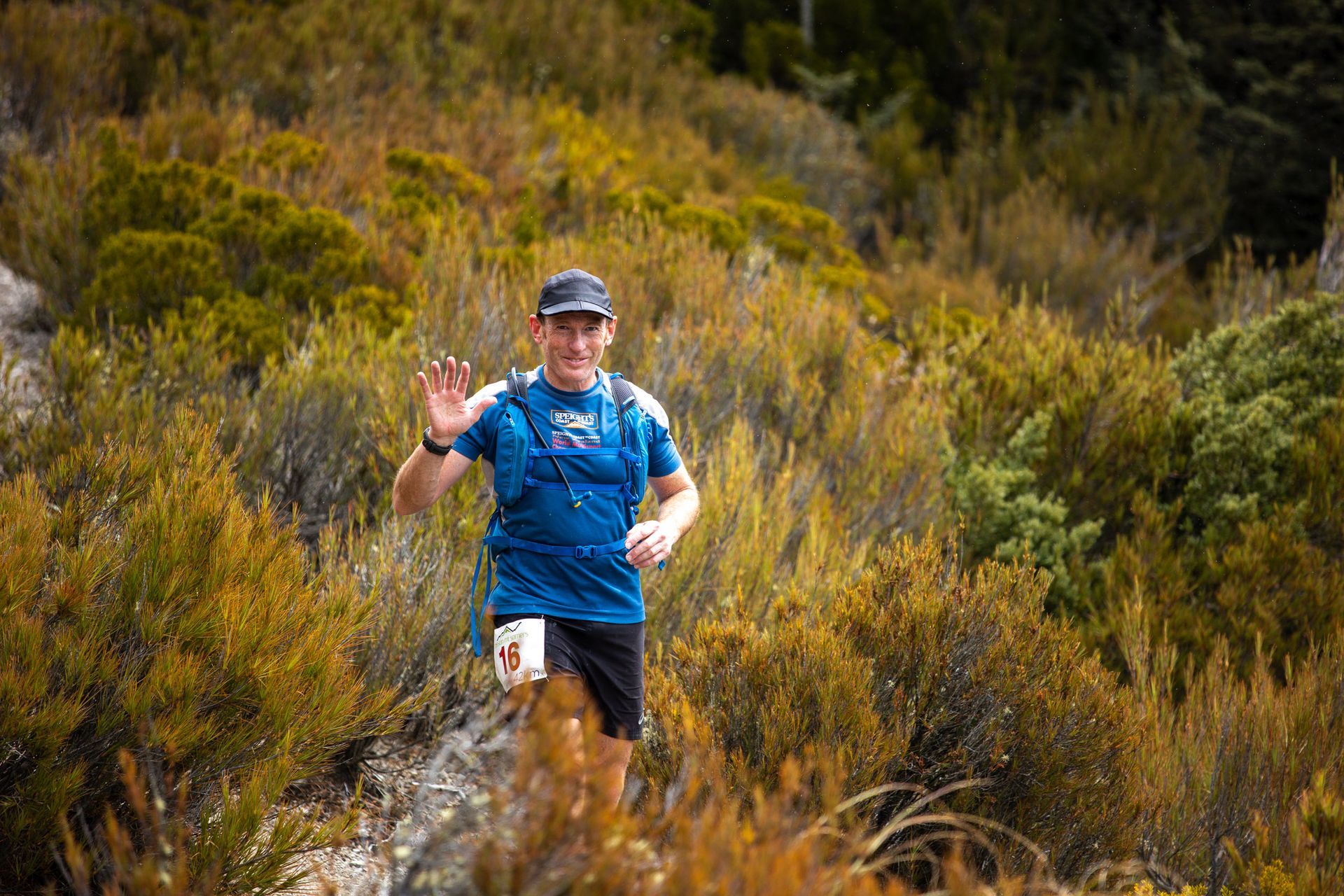 A man is running in the woods and waving at the camera.