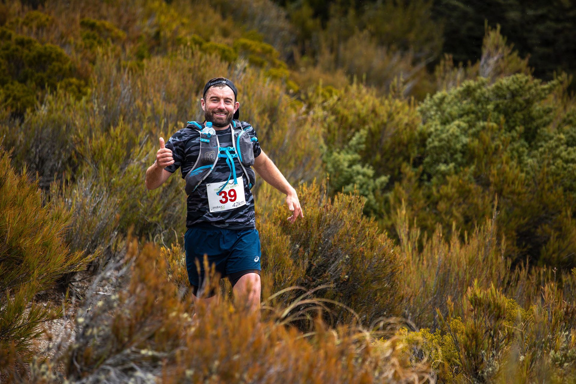 A man is running on a trail in the woods and giving a thumbs up.
