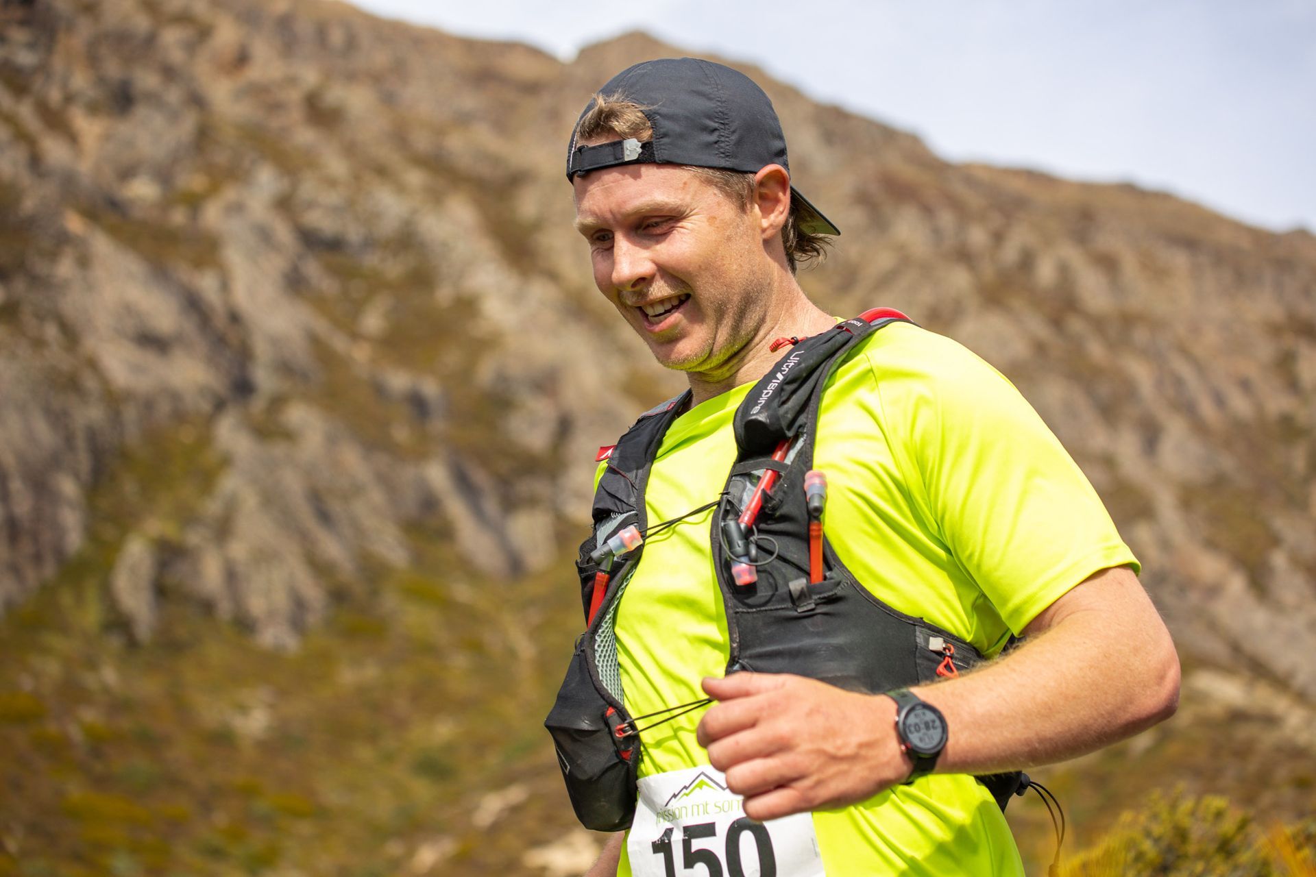 A man wearing a yellow shirt and a black hat is standing in front of a mountain.