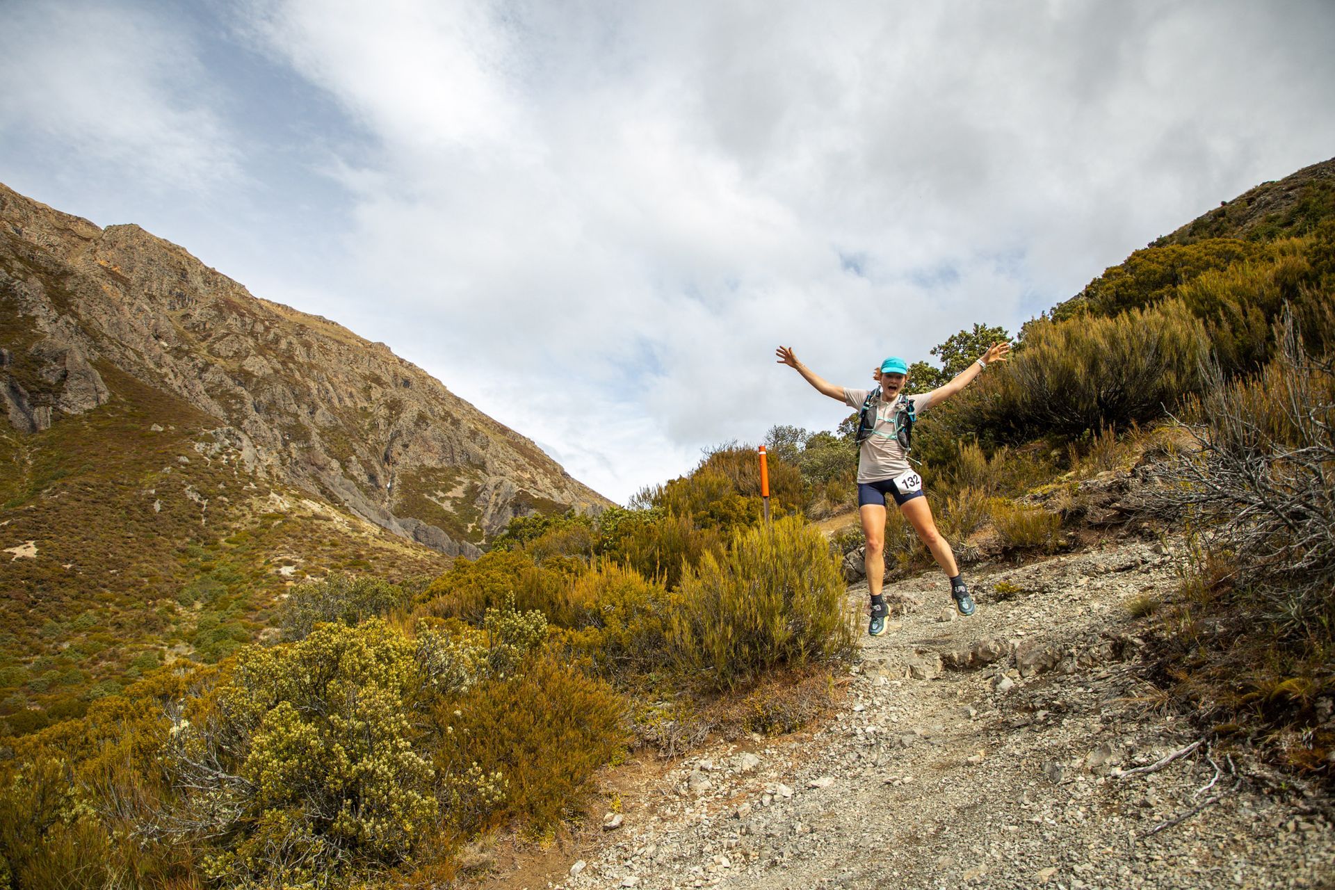 A woman is standing on a dirt path in the mountains with her arms outstretched.