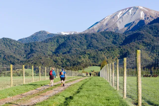 Two people are walking down a dirt road with mountains in the background.