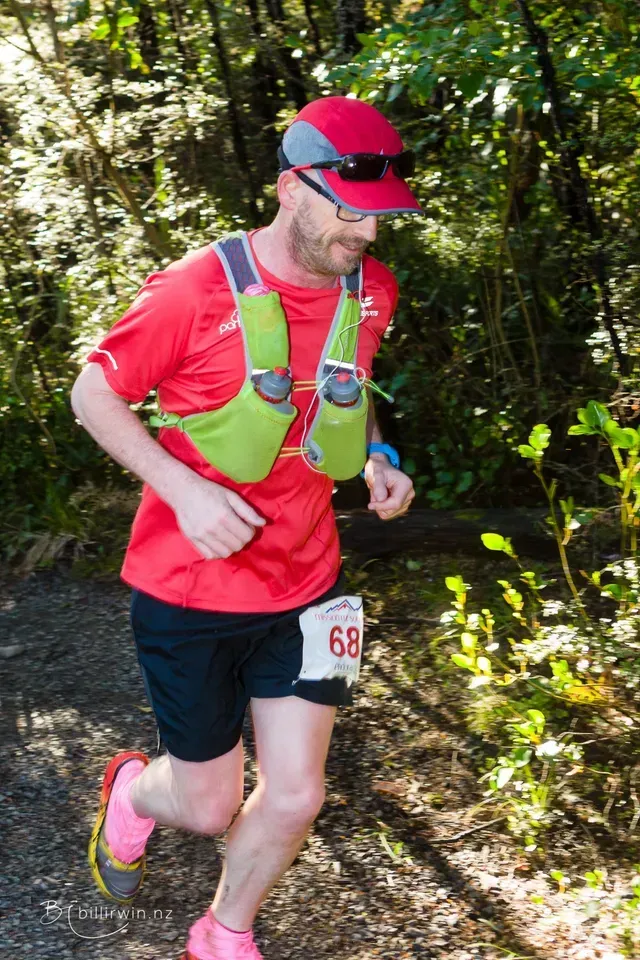 A man in a red shirt and green vest is running through the woods.