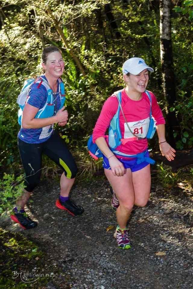 Two women are running on a trail in the woods.
